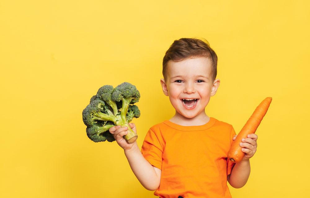 Junge vor gelben Hintergrund lacht fröhlich mit einem Brokkoli in der rechten und einer Möhre in der linken Hand