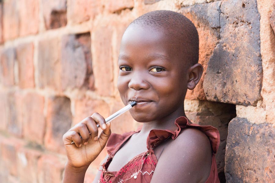 Afrikanisches Schulmädchen lehnt an einer Wand und hält lächelnd einen Kugelschreiber an die Lippen.
