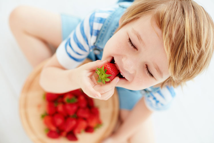 Ein Junge mit einer Schale Erdbeeren auf dem Schoß beisst genießerisch in eine Erdbeere.