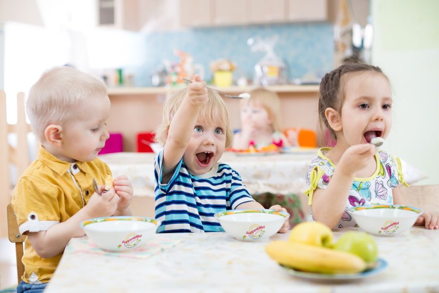 Drei Kinder essen gemeinsam in einer Kita. Der Junge hält freudig den Löffel mit Essen in die Höhe.