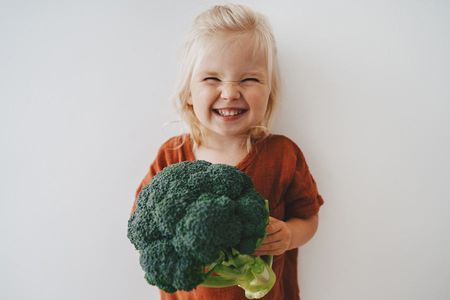 Ein kleines blondes Mädchen mit einem großen Brokkoli freut sich riesig und grinst mit zusammengekniffenen Augen.