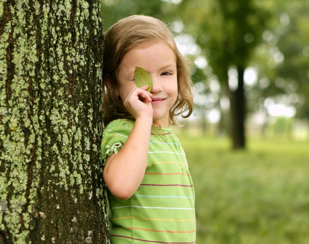Ein Mädchen mit grünem T-Shirt lehnt an einem Baumstamm und hält sich ein Blatt von einem Baum vor das rechte Auge.