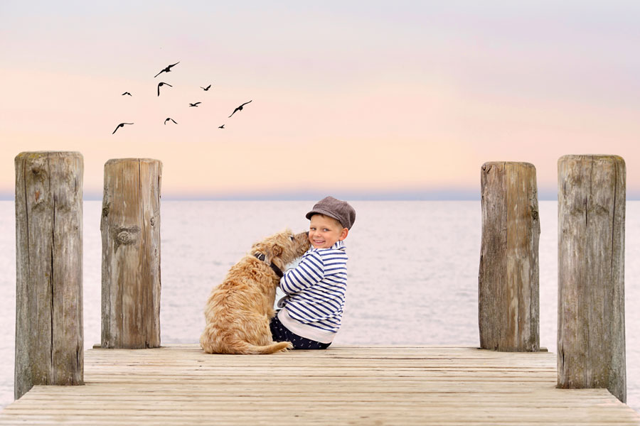 Ein kleiner Junge mit Schirmmütze und blau-weiß geringeltem Shirt sitzt am Meer auf einem Steg und wird von einem kleinem, beigen struppigen Hund abgeleckt.